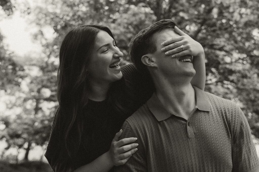 Bella laughing as she covers Joey’s eyes playfully while they stand together under the trees.