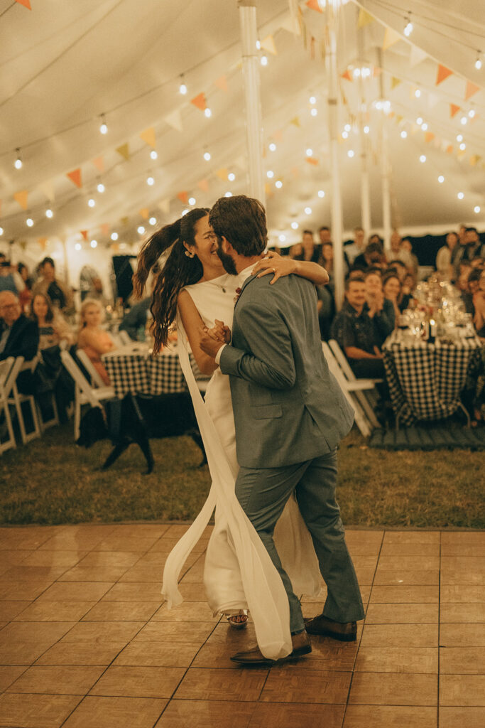 Eura and Stu sharing their first dance beneath a sailcloth tent strung with café lights, surrounded by guests — a warm, cinematic backyard wedding reception moment.