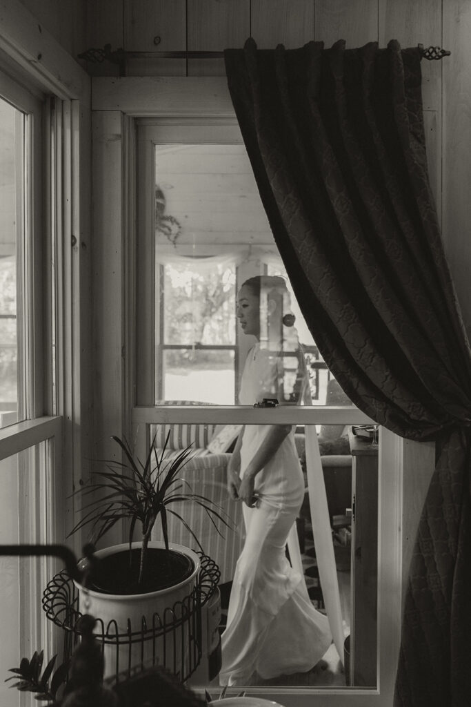 Eura standing on the farmhouse porch in her wedding dress, framed through the window as she prepares for her Minnesota backyard wedding ceremony.