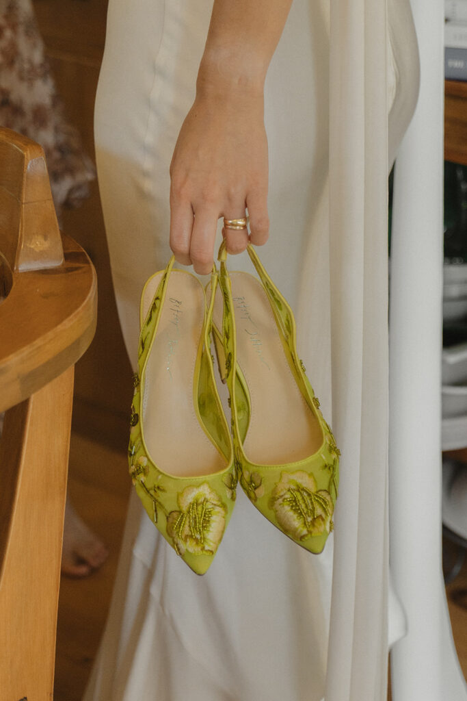 Eura holding her green embroidered wedding shoes on the morning of her backyard wedding, a playful pop of color for her garden-party style.