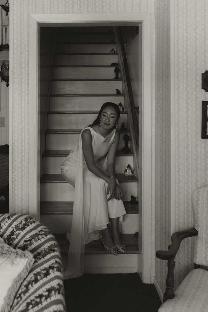 A candid black and white portrait of Eura sitting on the farmhouse staircase before her backyard wedding ceremony, full of quiet anticipation.