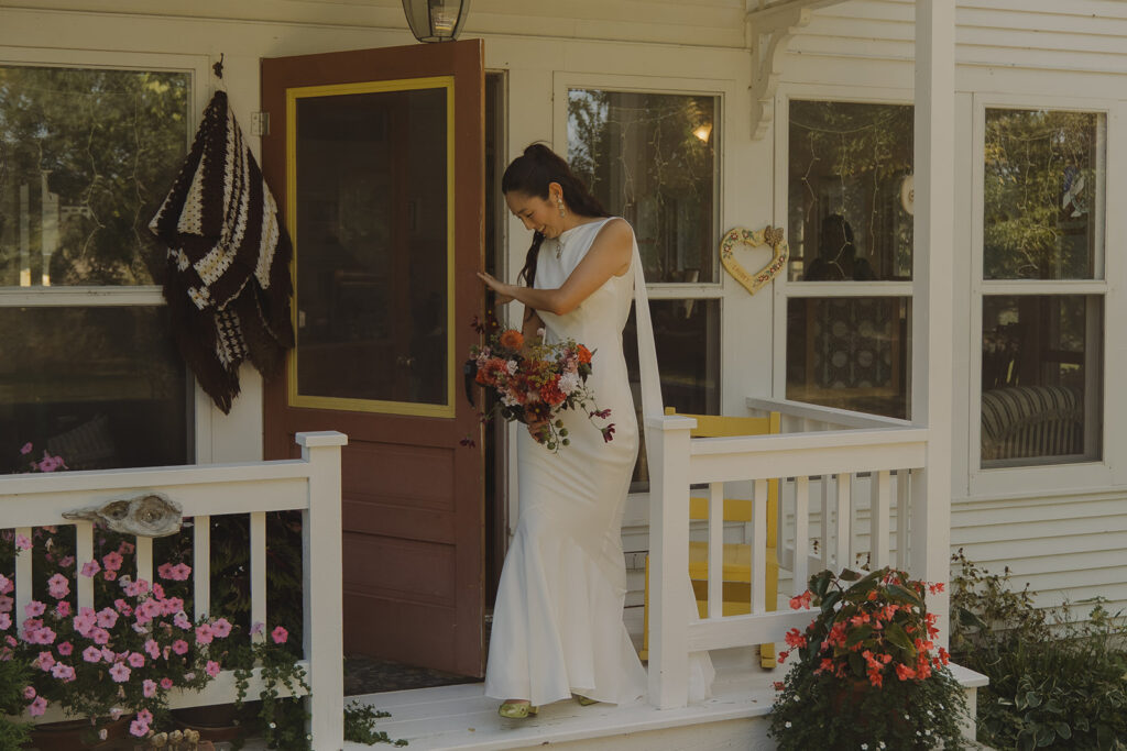 Eura standing on her grandma-in-law’s farmhouse porch with her bouquet before the backyard wedding ceremony, a quiet moment in the morning light.