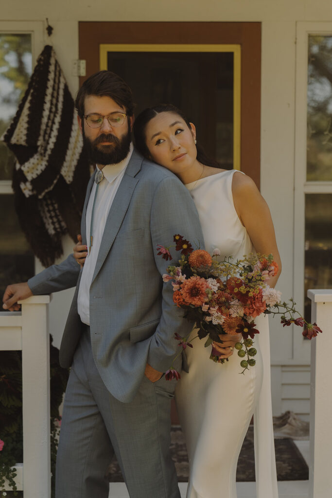 Eura and Stu leaning together on the porch of his grandma’s farmhouse during their backyard wedding, holding bright garden-party florals and showing how effortless a at-home celebration can look.