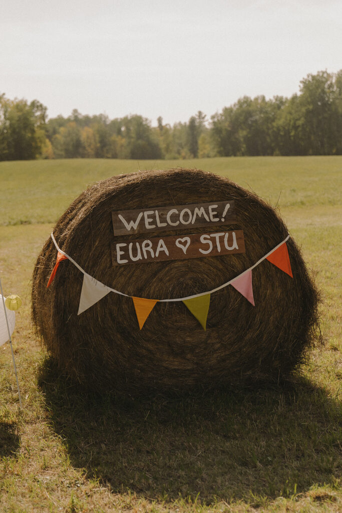 Hay bale welcome sign with handmade bunting at Eura and Stu’s backyard wedding, a charming garden wedding party detail on the farmhouse lawn.