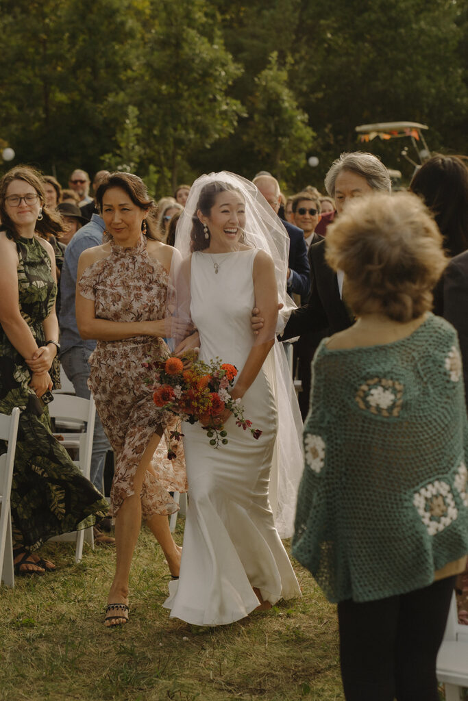 Eura walking down the aisle at her backyard wedding while holding colorful garden-party florals, greeted by loved ones.