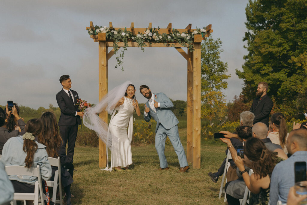 Eura and Stu making a playful ceremony exit under a wooden floral arch during their backyard wedding, surrounded by cheering guests on the hill.