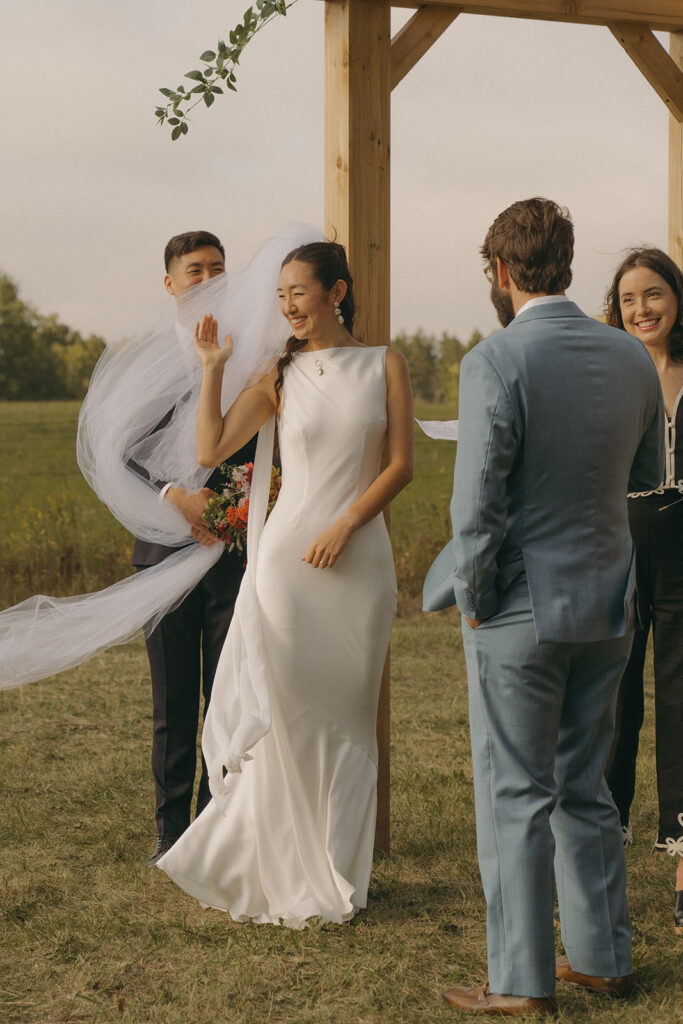 Wind lifting Eura’s veil during the ceremony as her brother steadies it, a candid moment from this outdoor backyard wedding.