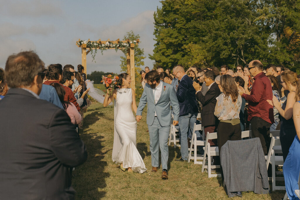Eura and Stu walking up the aisle hand-in-hand after their outdoor ceremony on the hill, surrounded by cheering guests — a joyful moment from their backyard wedding at Grandma’s house.