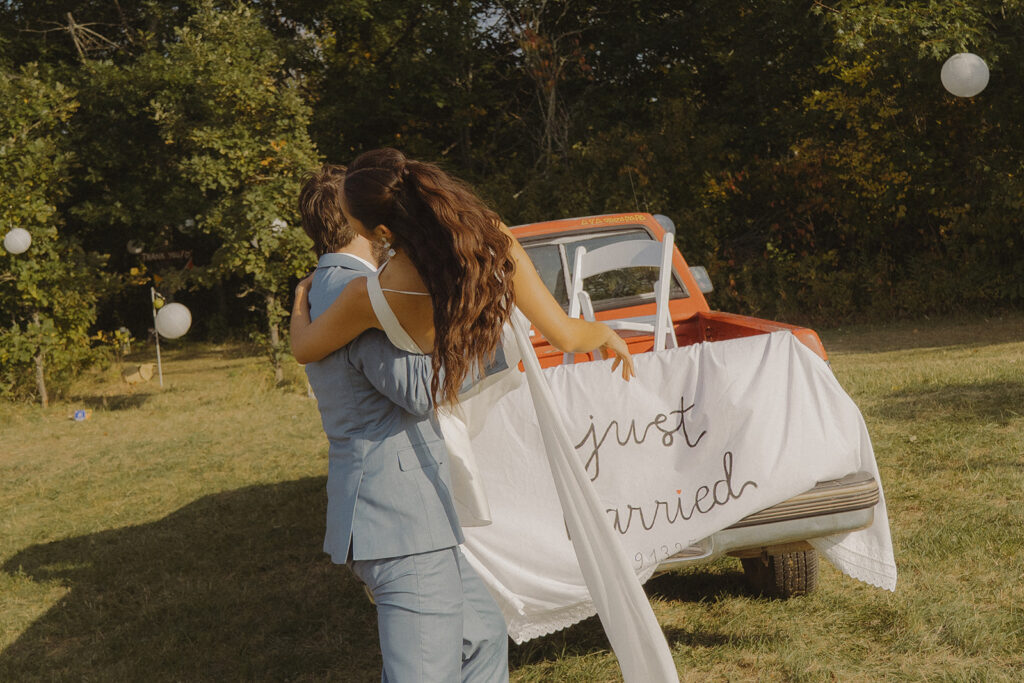 Eura and Stu hugging beside their vintage red “Just Married” truck in the backyard, a sweet and nostalgic end-of-ceremony moment from their farmhouse wedding celebration.