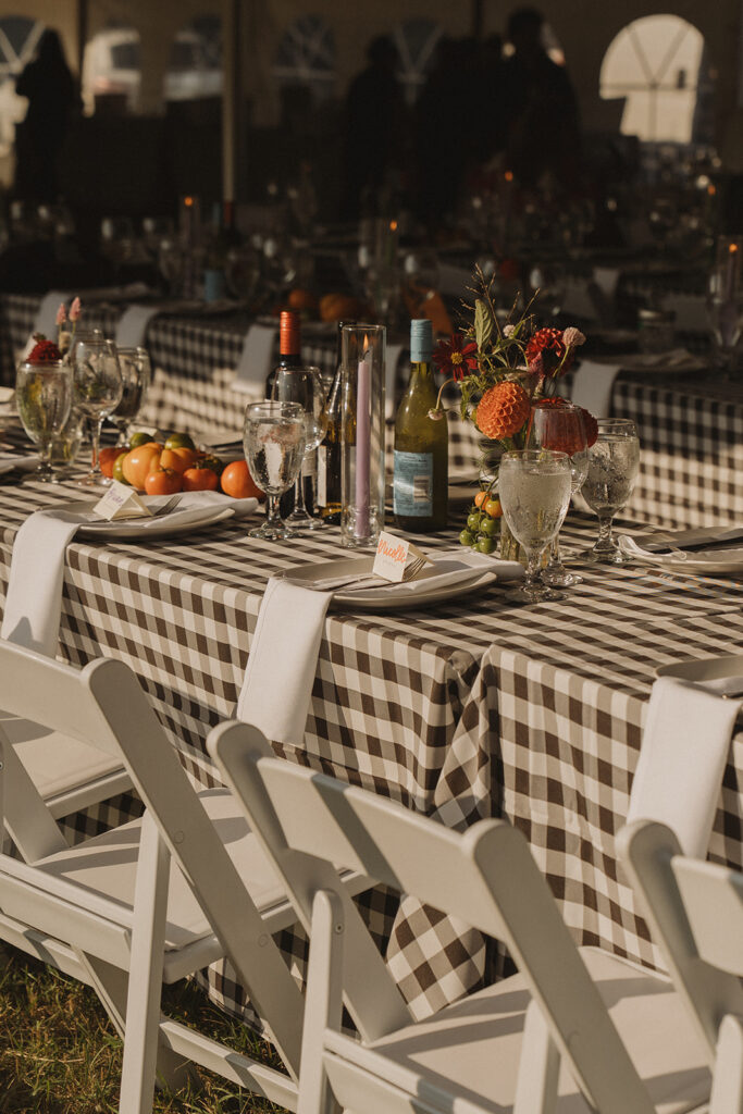 Long harvest tables set with gingham linens, candles, wine, and tomatoes at Eura and Stu’s backyard wedding — an elevated, homegrown reception look full of garden-party inspiration.