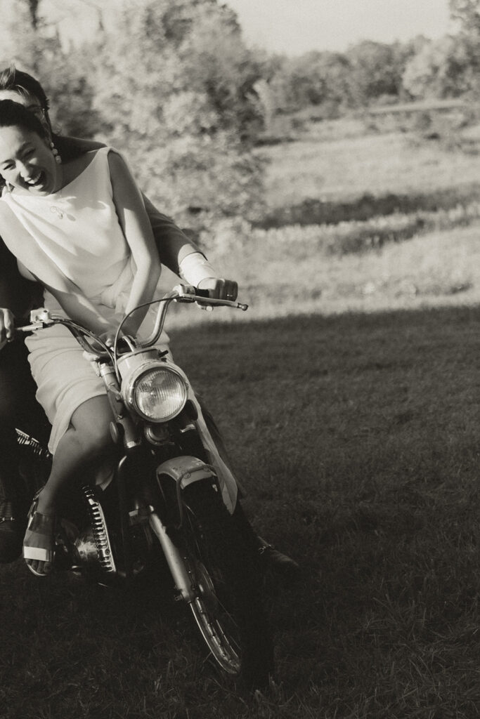 Black and white photo of Eura and Stu laughing on the dirt bike during their backyard wedding, capturing a candid and joyful moment.