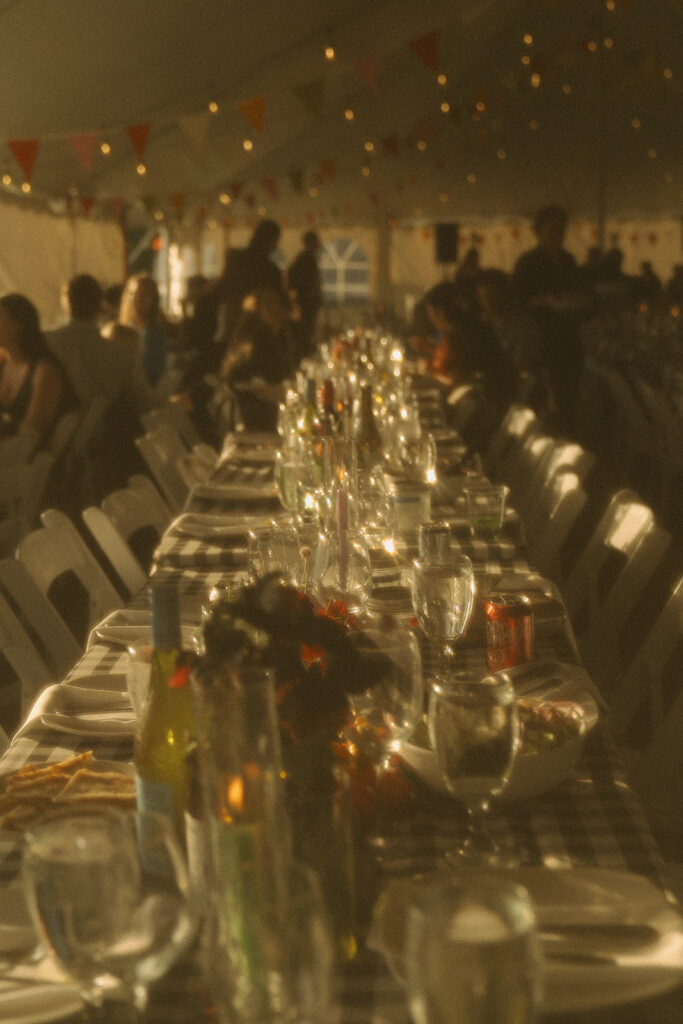 Long reception tables set with gingham linens and glowing candles under a sailcloth tent, creating a warm backyard wedding atmosphere.