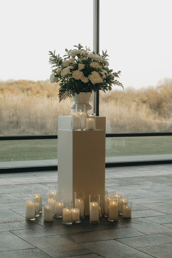 Candles in hurricane vases sit around a white pedestal topped with sprawling white roses