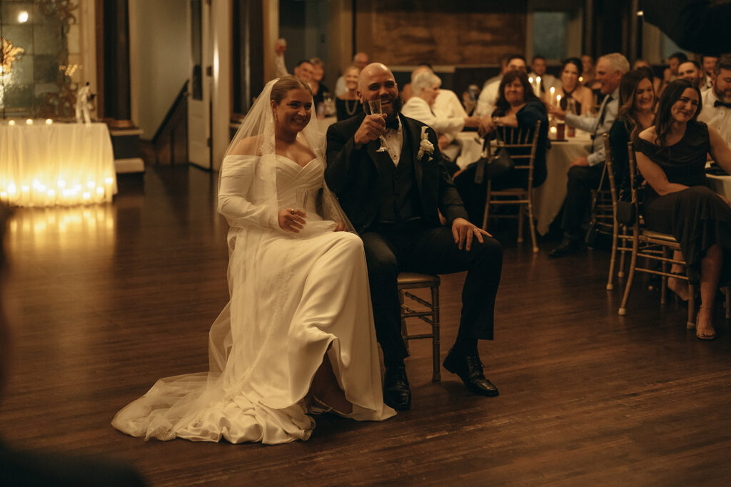 Groom toasts to person giving a speech as bride grins 