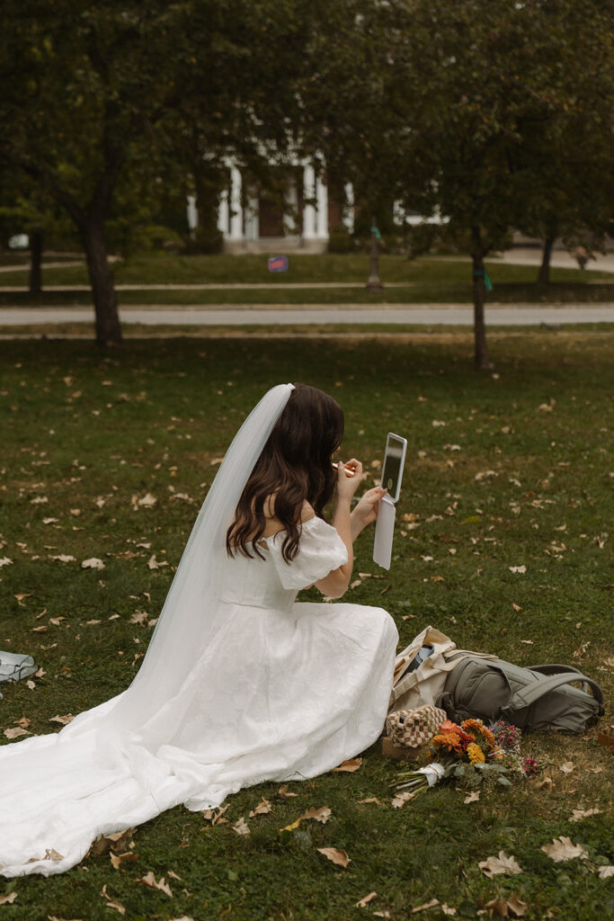 Vintage wedding bride sits down in the grass to touch up her make up in vintage wedding pics