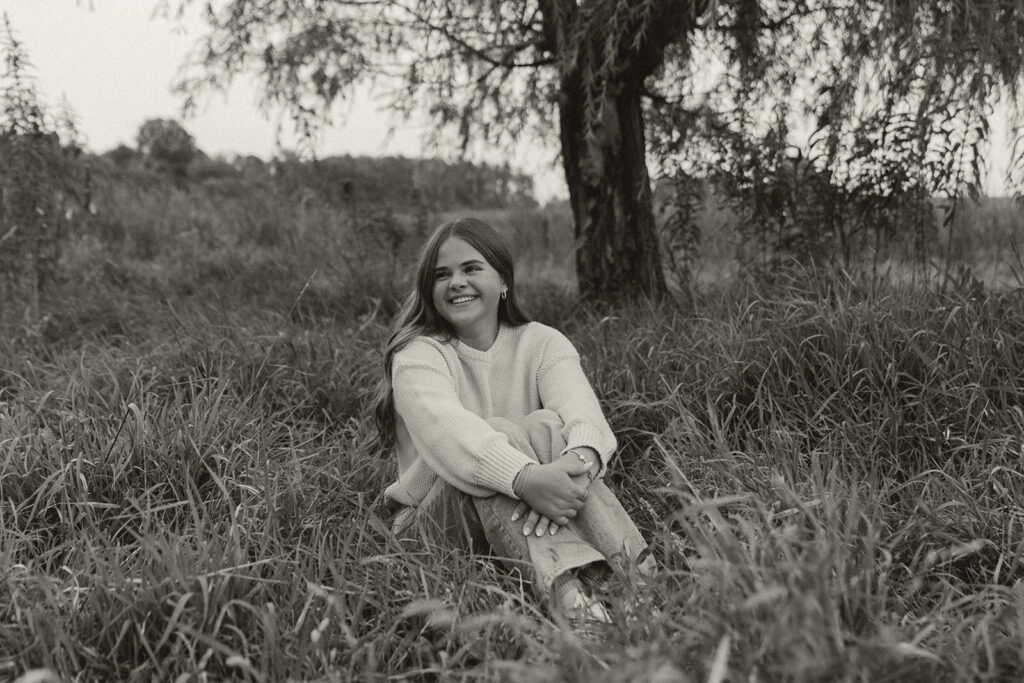 Young girl sits in meadow grass wearing white sweater and jeans for her senior photos
