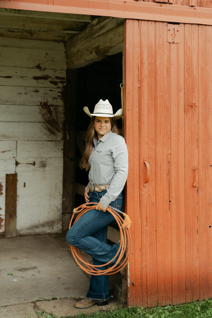 High school senior leans against red barn door while holding a red lasso and wearing a white cowboy hat