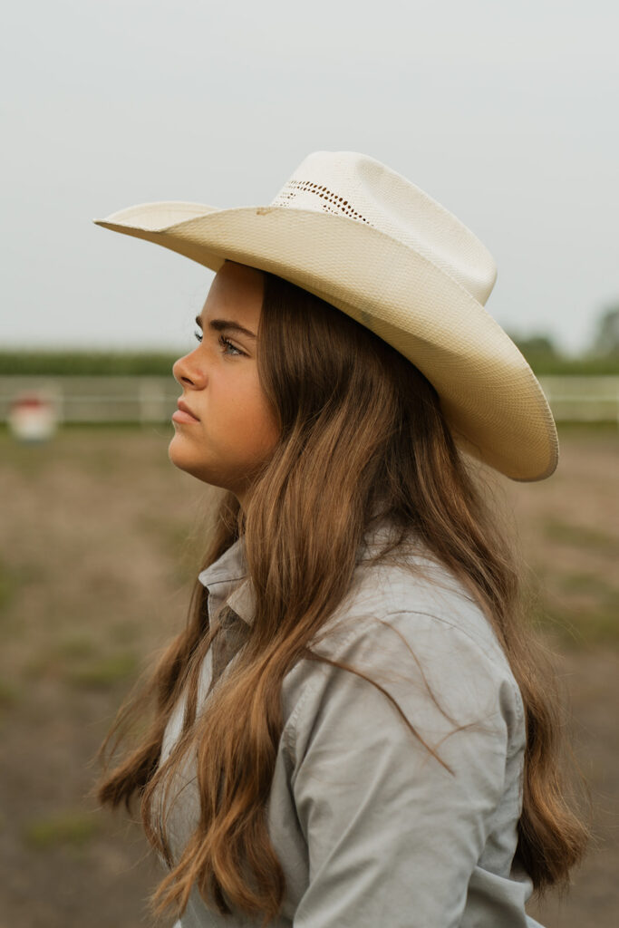 Girl with long, brown hair wearing a cowboy hat looks off in the distance as her profile is captured in her senior photos