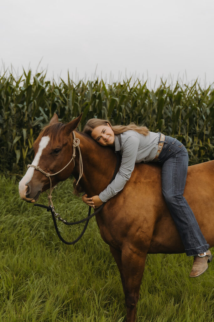 Girl in denim and a button down lays down on her horse and hugs her