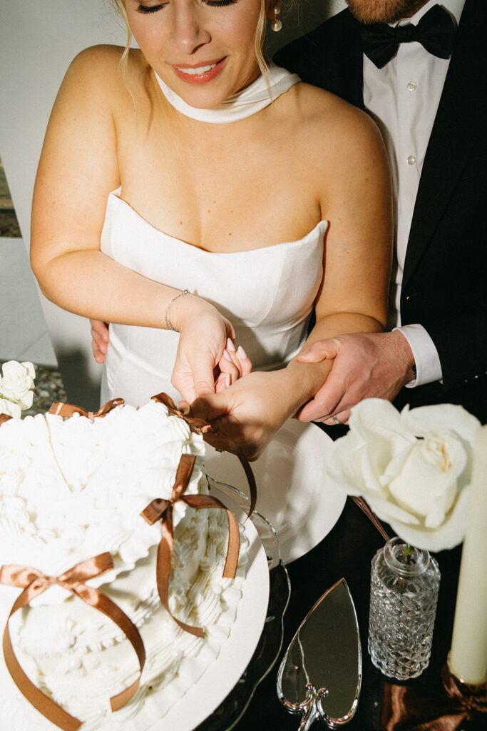 Groom holds bride's hand steady from behind as she cuts into their vintage cake during cake cutting at wedding
