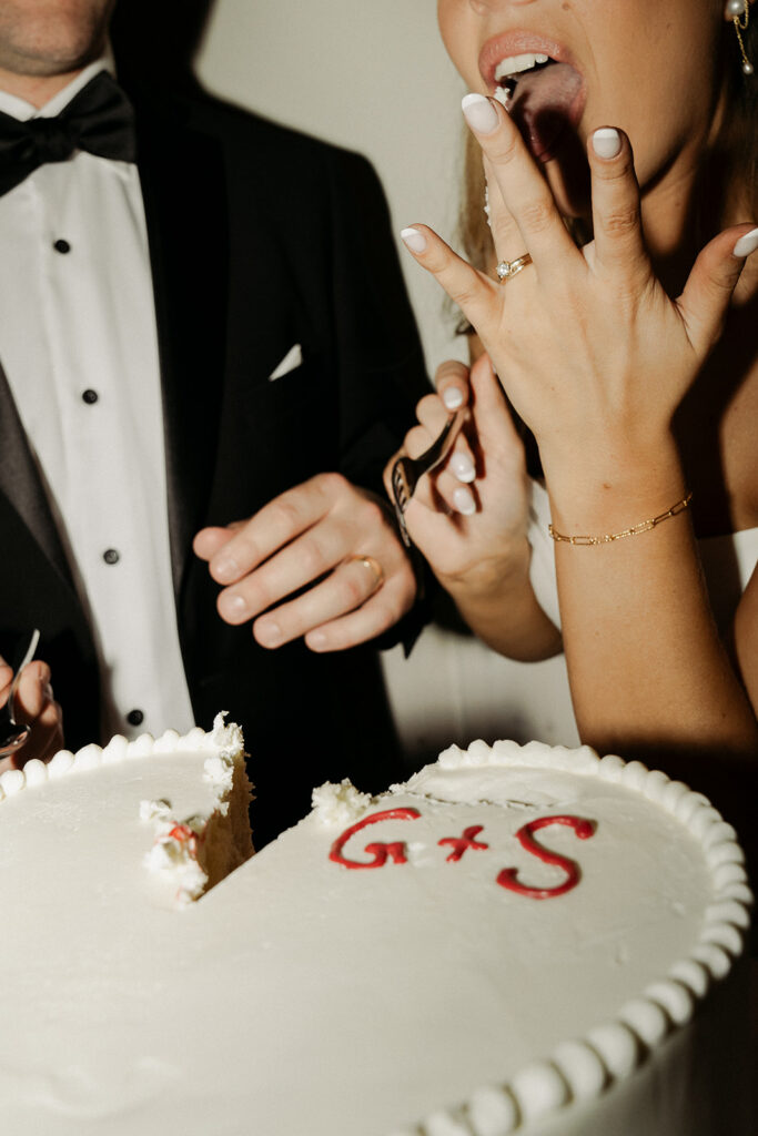 Heart-shaped vintage white wedding cake with G+S inscribed on the top with bright red frosting sits in the foreground while bride licks her frosting fingers in the background