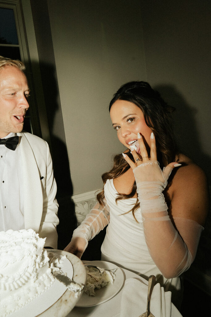 Bride playfully licks frosting off her fingers while groom acts scandalized for the bit in flirty wedding cake pictures
