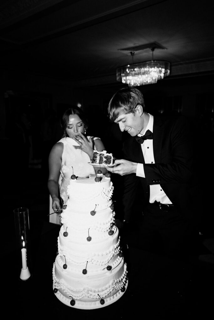 Groom balances a slice of cake on a plate while bride licks frosting from her fingers