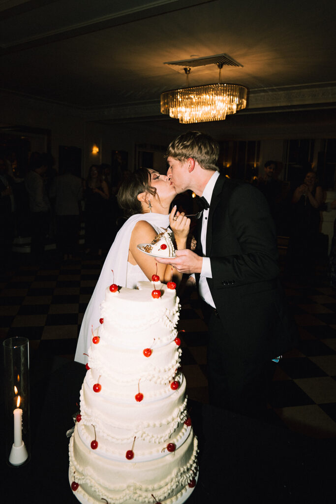 Vintage, art deco cake cutting pictures at wedding with bride and groom kissing and multi-tiered vintage wedding cake topped with cherries towering in the foreground.