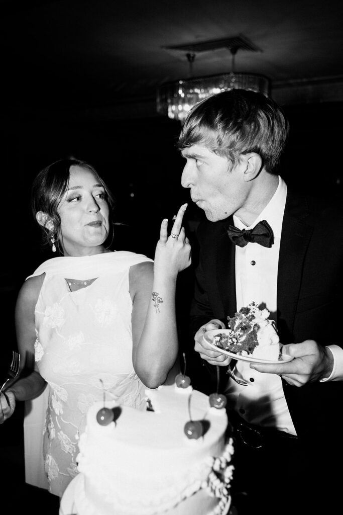 Bride playfully feeds groom some frosting from their vintage wedding cake with her ring finger in classic wedding cake pictures