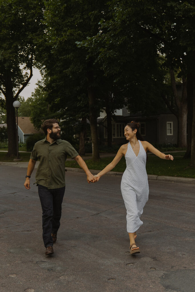 Couple walks hand in hand through a charming residential street, blending vintage engagement photos with warm Nancy Meyers aesthetic neighborhood romance.