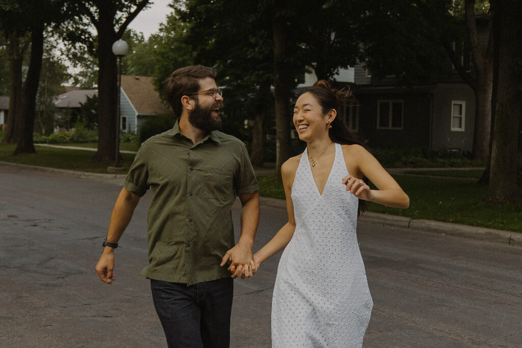 Couple runs hand in hand through the street at dusk, featuring playful 90s rom com chemistry and candid vintage engagement photos style.
