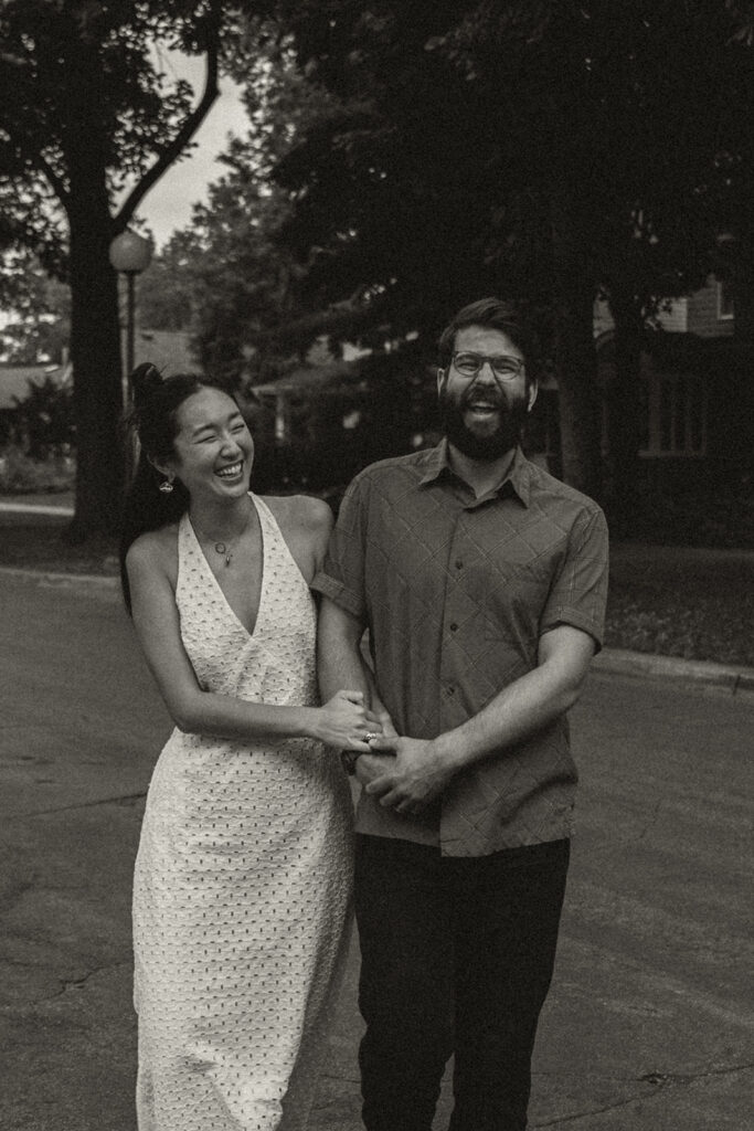 Black-and-white portrait of laughing couple walking down a tree-lined neighborhood street, capturing 90s rom com energy with vintage engagement photos charm.