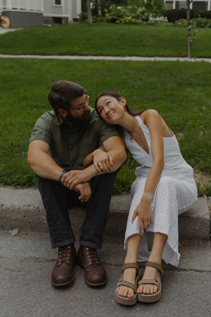 Couple sits on the curb smiling at each other in a quiet neighborhood, creating fun couple photoshoot ideas with a soft Nancy Meyers aesthetic.