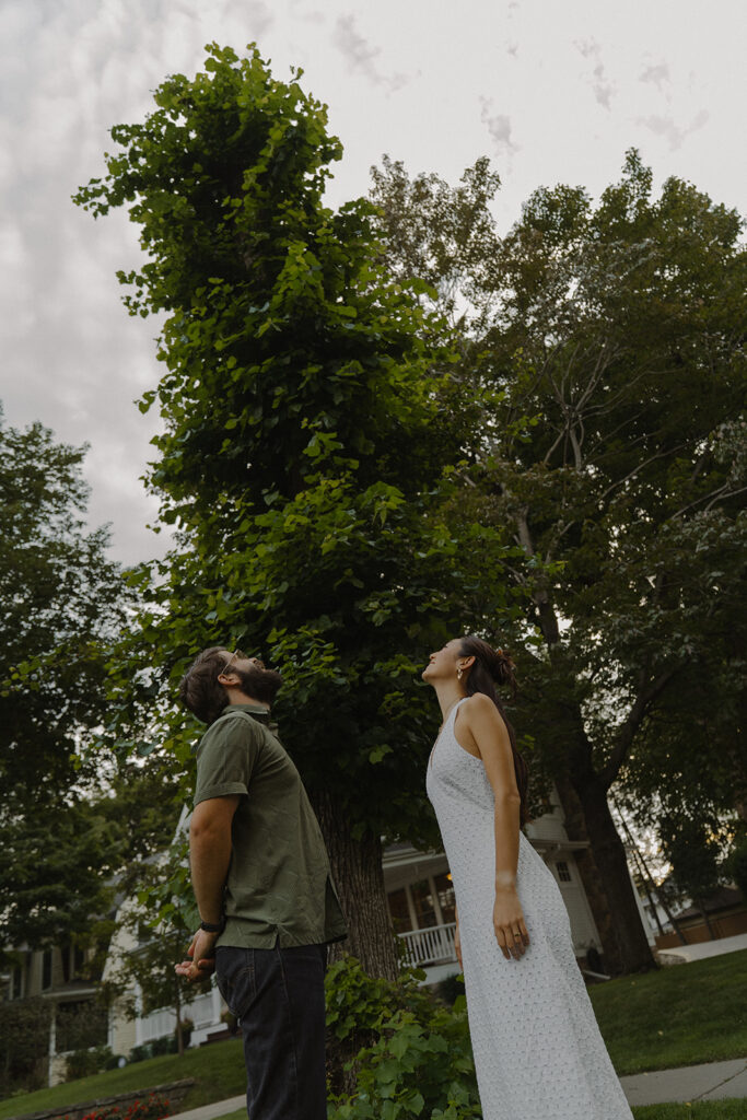 Couple stands beneath a towering ivy-covered tree looking upward, blending whimsical 90s rom com charm with vintage engagement photos style.