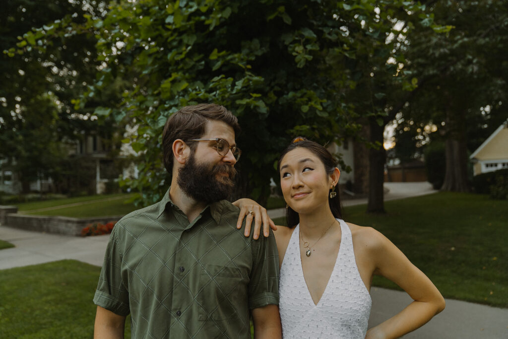 Couple smiles with playful side-eye in a leafy neighborhood street, capturing 90s rom com chemistry with candid vintage engagement photos energy.