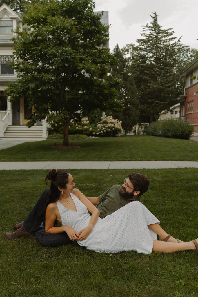 Couple lounges in the grass outside historic homes, creating fun couple photoshoot ideas with a soft Nancy Meyers aesthetic.