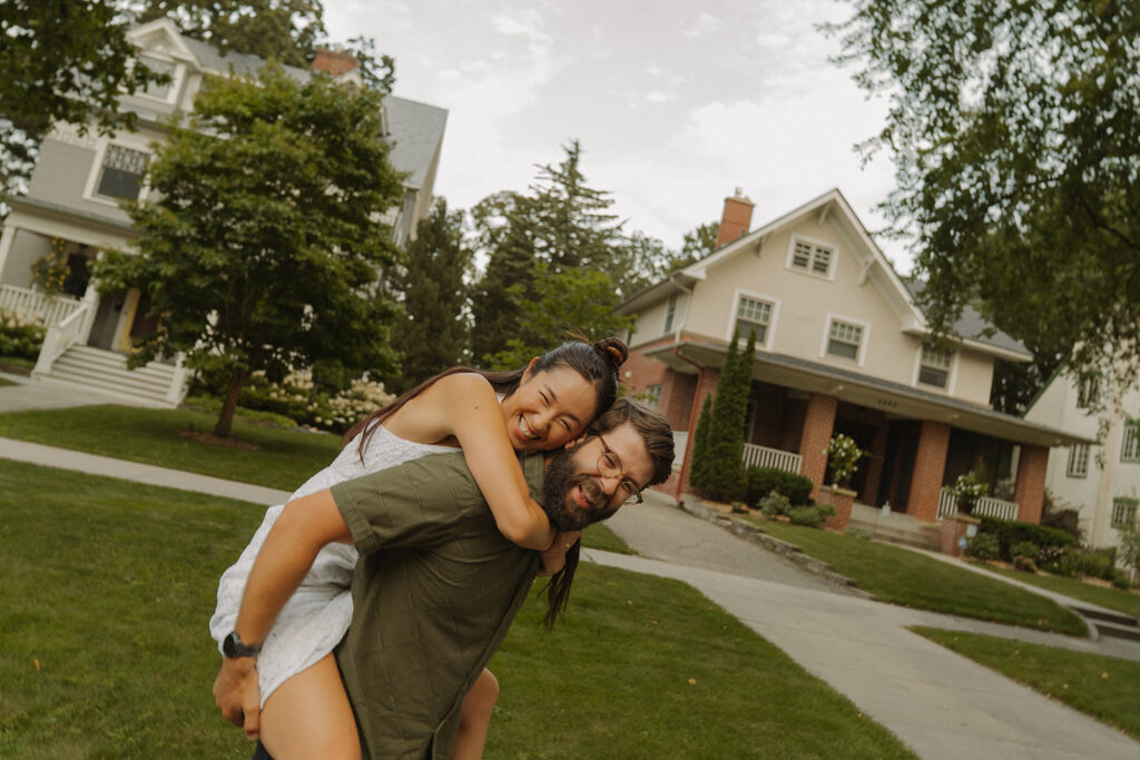 Groom gives bride-to-be a piggyback ride through a charming neighborhood lawn, featuring playful fun couple photoshoot ideas and nostalgic romance.