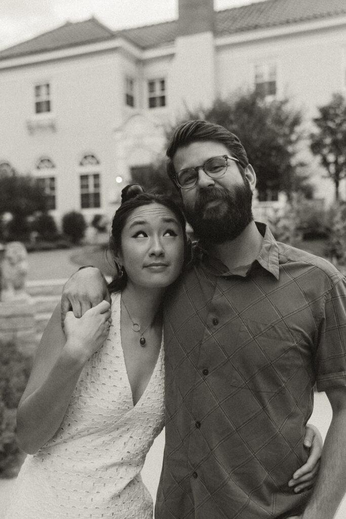 Black-and-white portrait of couple embracing in front of an elegant home, blending timeless vintage engagement photos with soft Nancy Meyers aesthetic style.