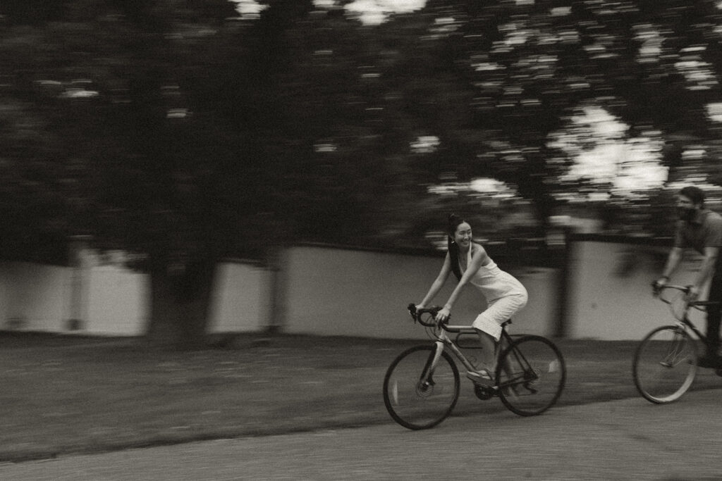 Couple rides bicycles through the neighborhood in motion blur, featuring cinematic 90s rom com movement and timeless vintage engagement photos vibes.