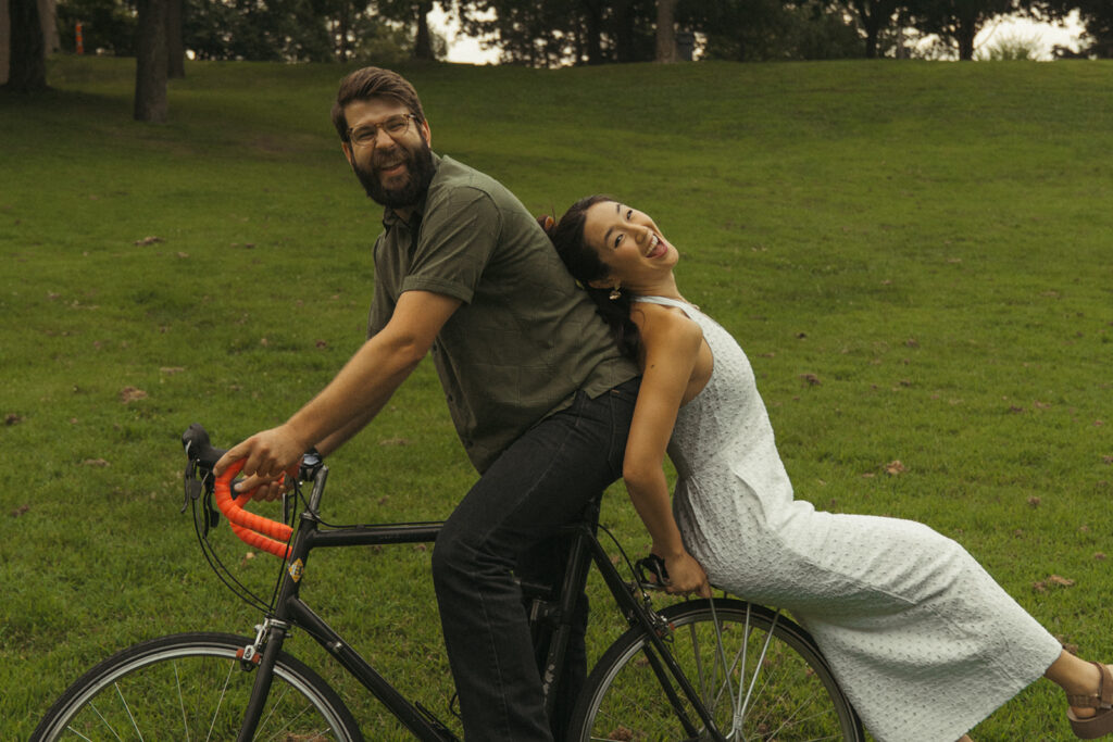 Couple poses back-to-back on a bicycle in the park, capturing playful 90s rom com energy with quirky vintage engagement photos charm.