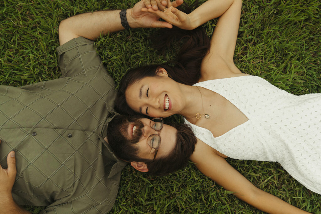 Couple lies in green grass smiling side by side, creating relaxed vintage engagement photos with dreamy 90s rom com energy.