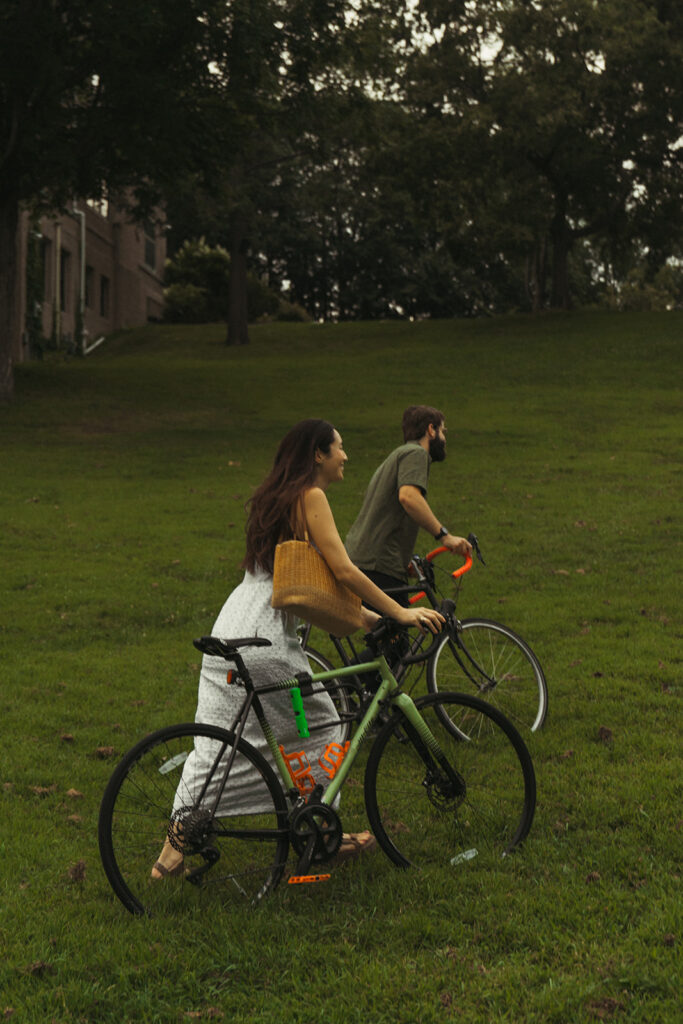 Couple walks their bikes uphill through a grassy park, blending relaxed Nancy Meyers aesthetic scenery with creative fun couple photoshoot ideas.
