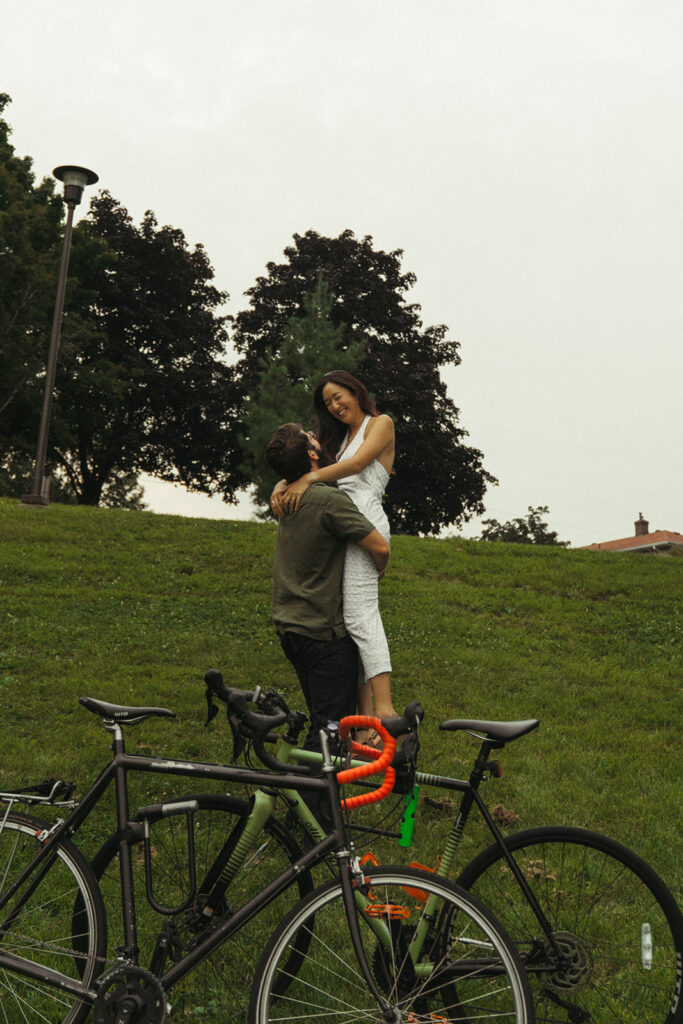 Groom lifts bride-to-be beside their bicycles on a hillside, capturing joyful 90s rom com romance and playful vintage engagement photos energy.