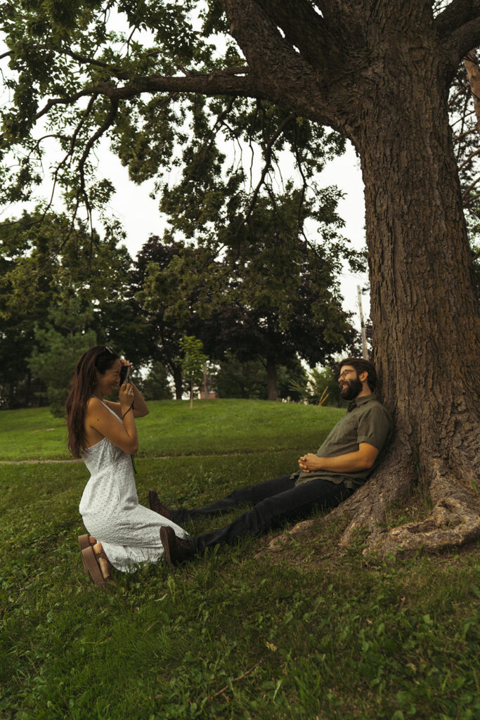 Couple kneels beneath a large tree while she photographs him with a film camera, capturing playful 90s rom com energy and vintage engagement photos charm.