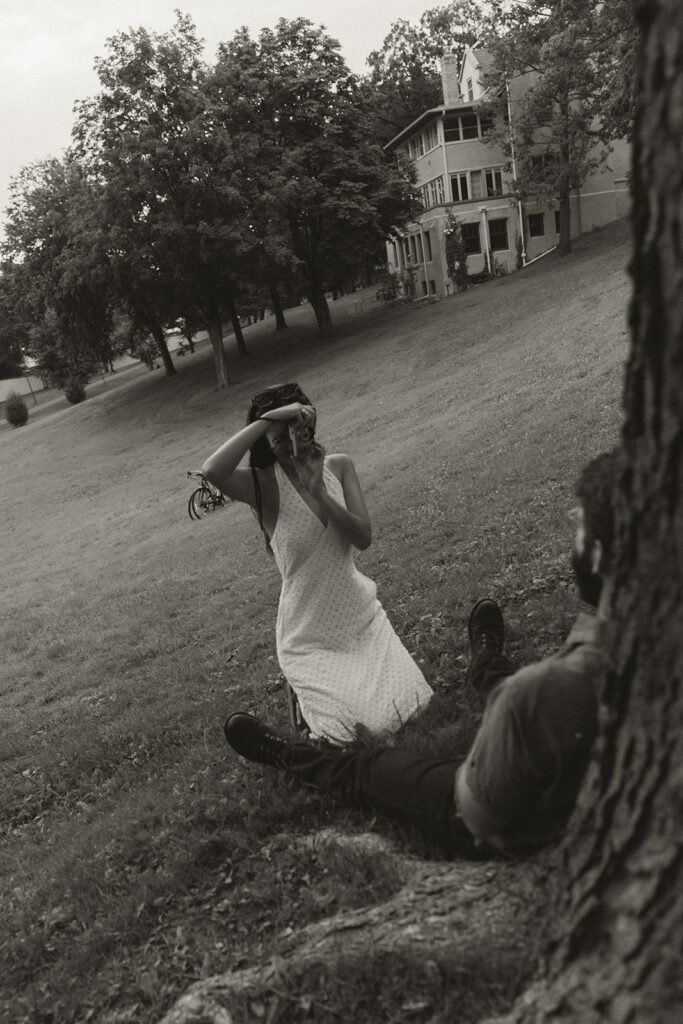 Black-and-white image of bride-to-be taking photos with a film camera while fiancé relaxes under a tree, creating candid fun couple photoshoot ideas with nostalgic style.