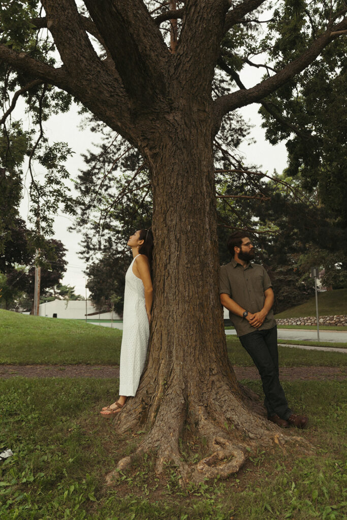 Couple poses on opposite sides of a large tree in a park, offering creative fun couple photoshoot ideas with playful storybook charm.