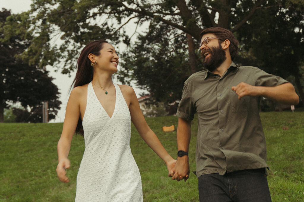 Laughing couple runs downhill holding hands in the park, featuring carefree 90s rom com chemistry and fun couple photoshoot ideas.
