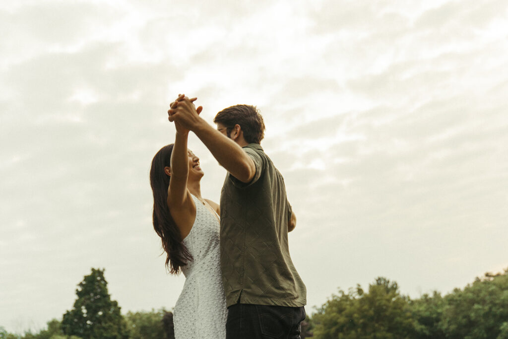 Couple slow dances outdoors beneath a cloudy sky, creating dreamy vintage engagement photos with romantic Nancy Meyers aesthetic warmth.