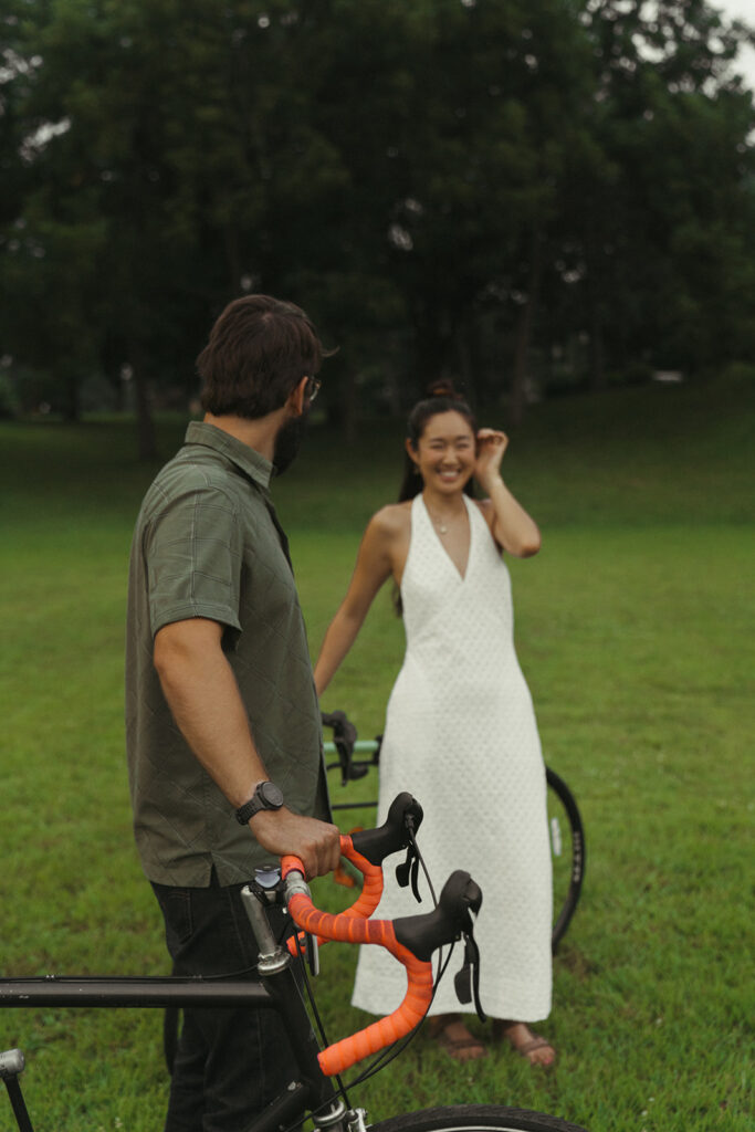 Groom stands beside bicycle smiling at bride-to-be in the grass, blending candid vintage engagement photos with a soft Nancy Meyers aesthetic.