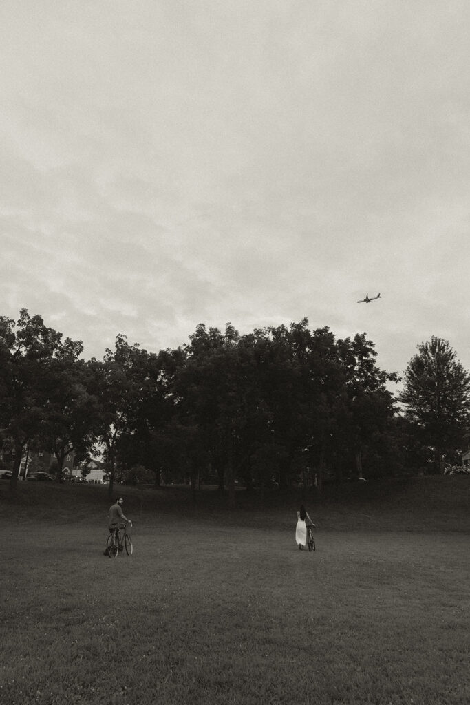 Wide black-and-white portrait of couple across an open park field with bikes nearby, inspired by spacious Nancy Meyers aesthetic storytelling and nostalgic romance.